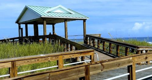 A wooden boardwalk leads to a gazebo overlooking the ocean, surrounded by tall grass and a clear blue sky.