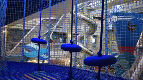 A colorful indoor play area featuring a netted climbing structure and a large slide in the background.