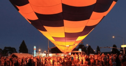 A crowd gathers under a brightly lit hot air balloon at dusk, with vibrant orange and black patterns.