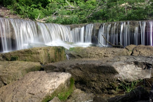 A serene waterfall cascades over rocky ledges, surrounded by lush greenery and natural scenery.