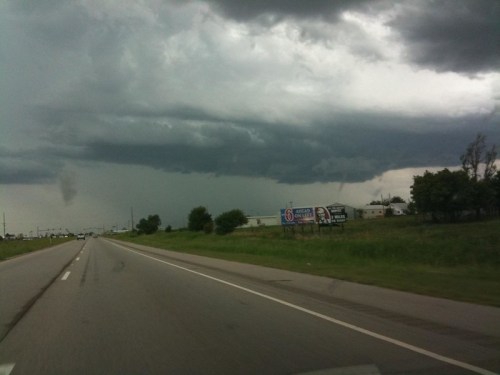 Dark storm clouds loom over a highway, with a billboard visible on the side and trees lining the road.