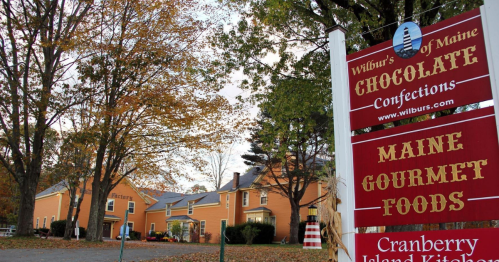 Sign for Wilbur's of Maine Chocolate Confections, surrounded by trees and autumn foliage, with a cozy building in the background.