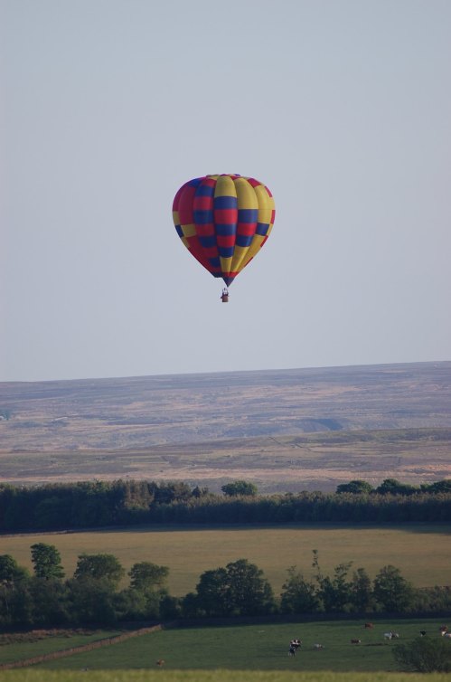 A colorful hot air balloon floats above a green landscape with distant hills under a clear blue sky.