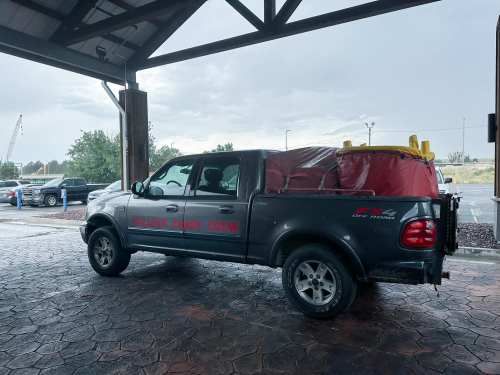 A black pickup truck with a red cover parked under a shelter, with rain in the background and a construction crane visible.
