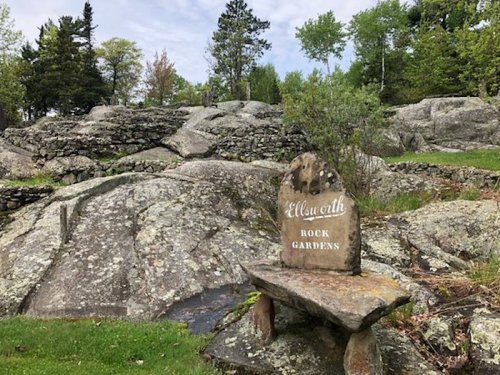 A stone bench in a rocky garden area, with a sign reading "Ellsworth Rock Gardens" surrounded by greenery.