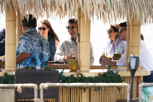 A group of people enjoying drinks at a tiki bar, wearing sunglasses and casual attire, with a tropical setting.