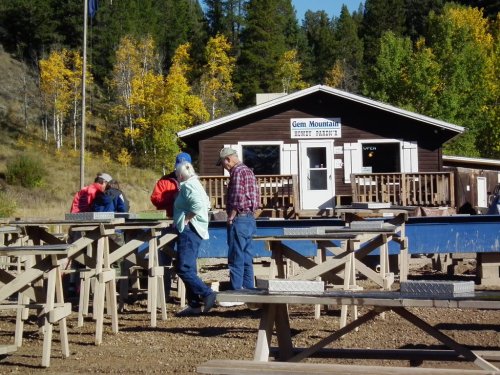 A group of people stands near wooden tables in front of a rustic building surrounded by trees in a mountainous area.