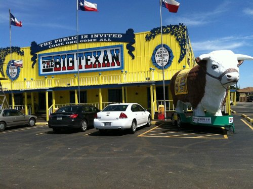 A large yellow building with "The Big Texan" sign, featuring a giant cow statue outside and parked cars nearby.