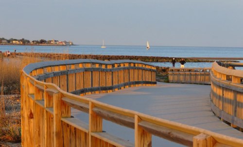 A wooden boardwalk curves along the shore, with sailboats in the distance and soft evening light reflecting on the water.