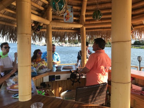A group of people socializing at a tiki bar by the water, with one person at the bar's helm.