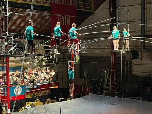 A circus performance featuring acrobats on a high wire, with one hanging and others on bicycles, in front of an audience.