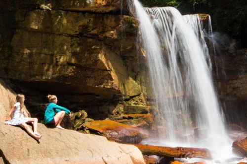 Two girls sit on rocks by a waterfall, enjoying the serene natural surroundings on a sunny day.