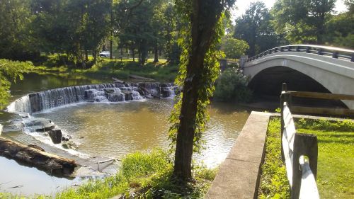A serene scene featuring a small waterfall beside a river, with a bridge in the background and lush greenery surrounding.