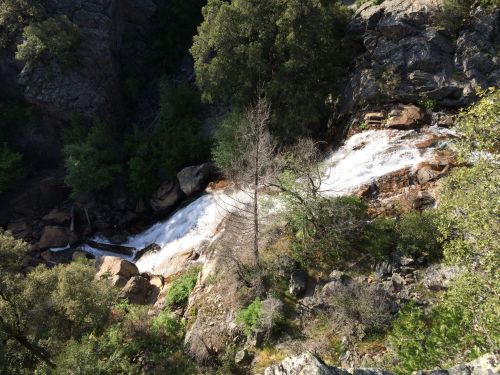 A cascading waterfall flows over rocks, surrounded by lush greenery and trees in a mountainous landscape.