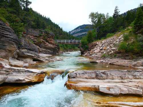 A serene river flows through rocky terrain, with a wooden bridge in the background and lush green trees lining the banks.