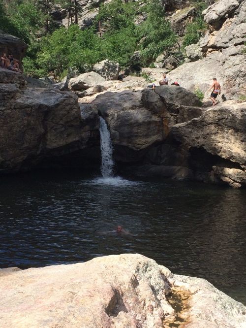 A natural swimming hole with a small waterfall, surrounded by rocky terrain and people enjoying the outdoors.