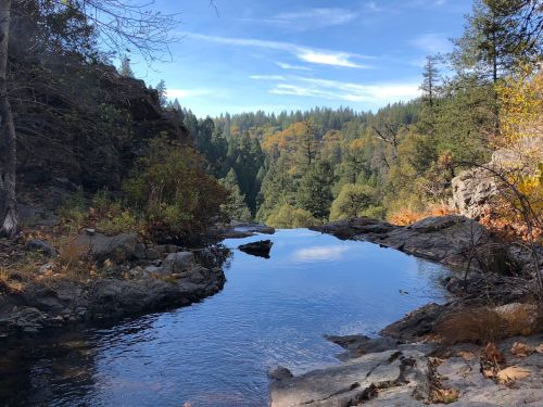 A serene river flows through rocky terrain, surrounded by trees and autumn foliage under a clear blue sky.