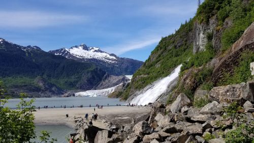 A scenic view of a glacier, waterfall, and mountains with people walking along a rocky beach.
