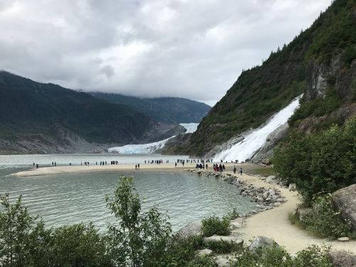A scenic view of a glacier with people walking along a sandy shore, surrounded by mountains and cloudy skies.