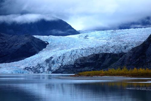 A serene landscape featuring a glacier reflecting in calm waters, surrounded by mountains and cloudy skies.