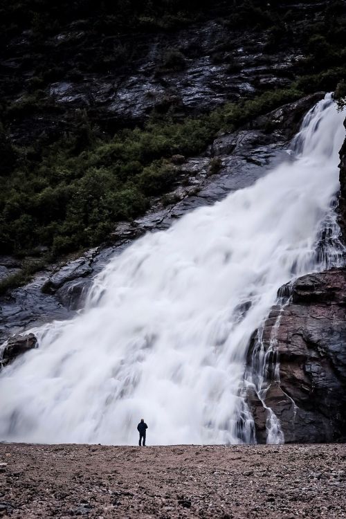 A person stands near a cascading waterfall, surrounded by rocky terrain and lush greenery.