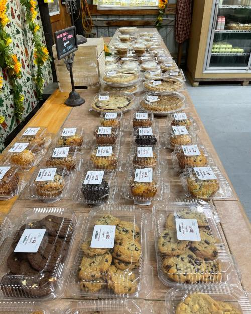 A display of various baked goods in clear containers, including cookies and pies, arranged on a wooden table.