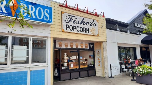 Exterior of Fisher's Popcorn shop with a sign, showcasing popcorn containers and nearby seating areas.