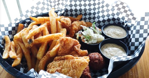 A basket of crispy fried fish, golden fries, coleslaw, and two dipping sauces on a checkered paper liner.
