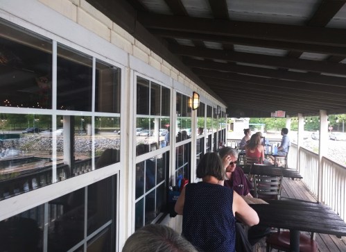 A view of a restaurant patio with diners seated at tables, overlooking a waterfront scene through large windows.