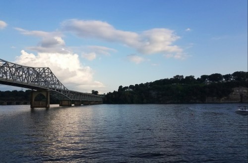 A bridge spans a calm river under a blue sky with clouds, surrounded by green hills and trees.
