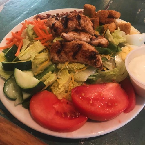 A colorful salad with grilled chicken, cucumbers, tomatoes, carrots, and croutons, served with a side of dressing.