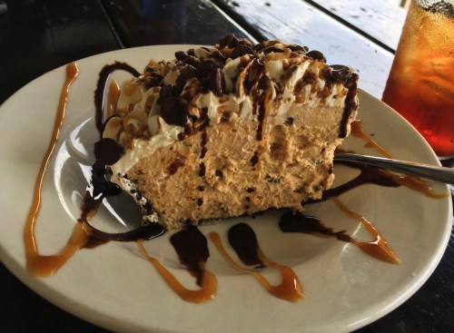 A slice of creamy dessert topped with whipped cream, chocolate drizzle, and nuts, served on a white plate.