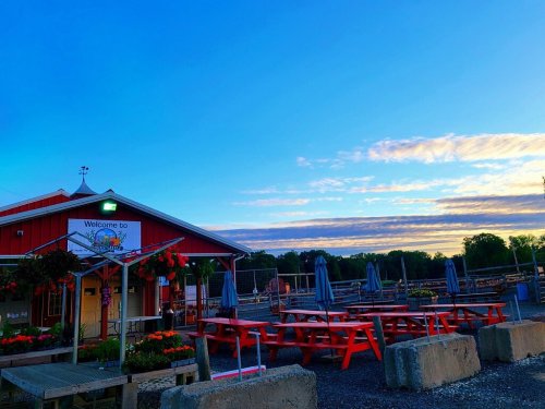 A red building with a welcome sign, surrounded by picnic tables and umbrellas, under a blue sky with clouds at sunset.