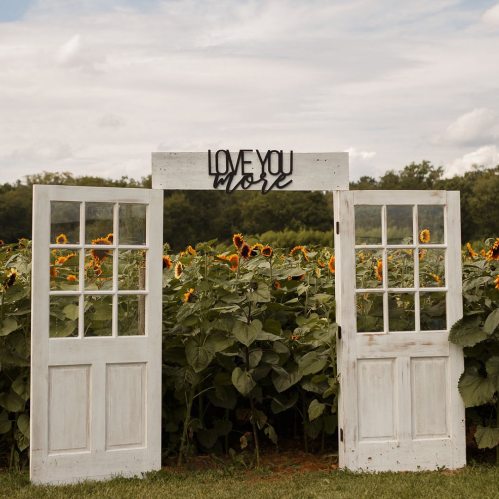 A rustic arch made of two white doors with a "Love You More" sign, surrounded by vibrant sunflowers under a cloudy sky.
