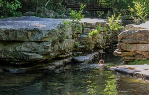 A person swims in a serene, rocky creek surrounded by lush greenery and a wooden bridge in the background.