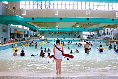 A lifeguard stands by a busy indoor pool filled with people enjoying the water and floating on tubes.