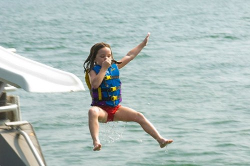 A child in a life jacket jumps off a slide into the water, splashing as they leap.