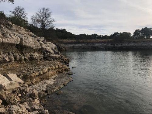 A calm body of water bordered by rocky shores and sparse trees under a cloudy sky.