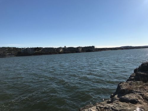 A calm body of water with a rocky shore and buildings visible in the distance under a clear blue sky.
