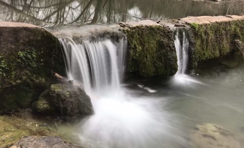 A serene waterfall cascades over moss-covered rocks into a calm pool, surrounded by trees and natural scenery.