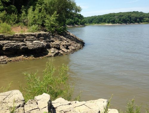 A serene riverbank scene with rocky shorelines, lush green trees, and clear blue skies reflecting on the water.