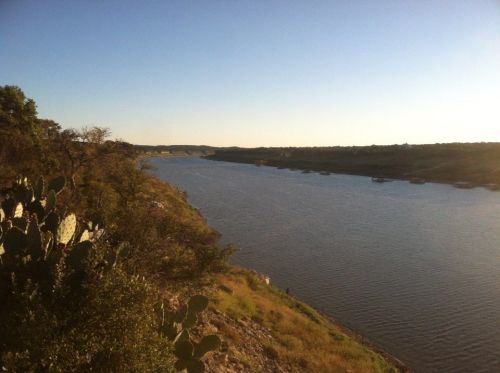 A serene river flows between lush greenery under a clear blue sky, with hills in the distance.