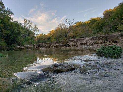 A serene river scene with clear water, rocky banks, and lush greenery under a blue sky with scattered clouds.