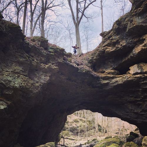 A person stands on a rocky ledge in a forest, surrounded by trees and a natural rock formation.