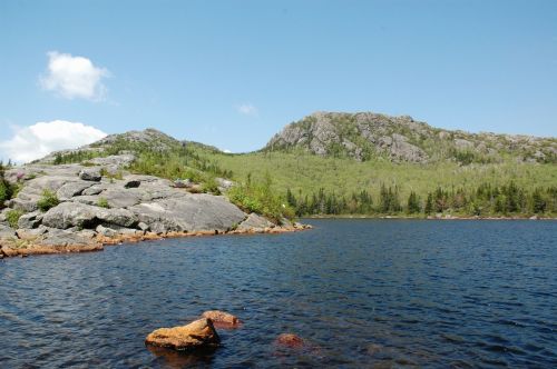 A serene lake surrounded by rocky shores and lush green hills under a clear blue sky.