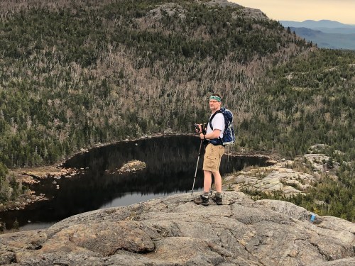 A hiker stands on a rocky outcrop, overlooking a forested landscape and a calm lake below.