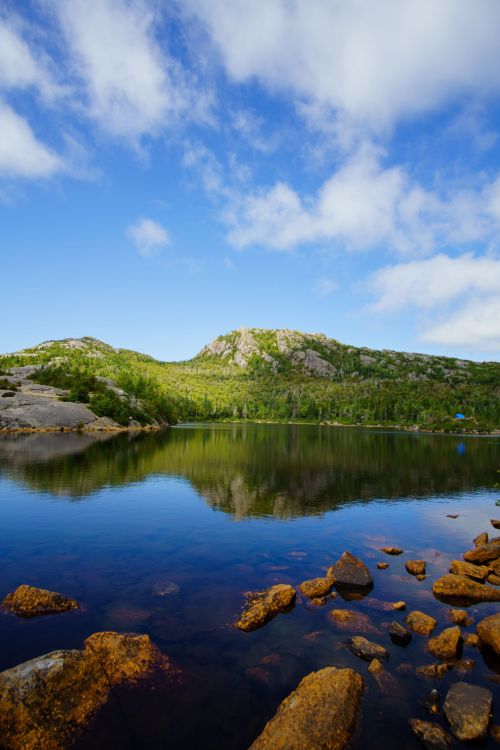A serene lake surrounded by green hills and rocky shores under a blue sky with fluffy clouds.
