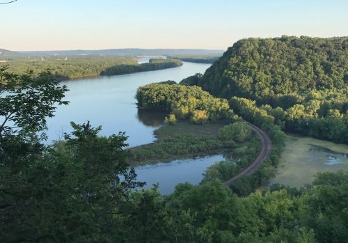 A scenic view of a winding river surrounded by lush green trees and hills under a clear blue sky.