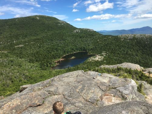 A scenic view from a rocky summit overlooking a forested landscape and a small lake under a blue sky.