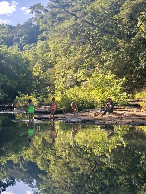 Four people stand by a serene riverbank, surrounded by lush greenery, with their reflections visible in the water.